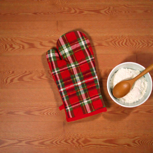 Guanto da cucina Holly in plaid a tema netalizio su un piano in legno con vicino una ciotola con farina e cucchiaio in legno.